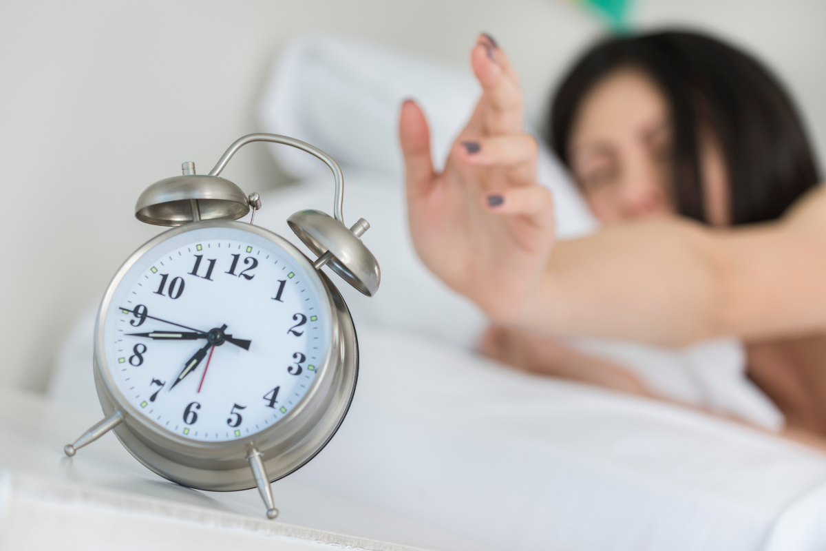 picture of a woman in bed reaching out to snooze her alarm clock